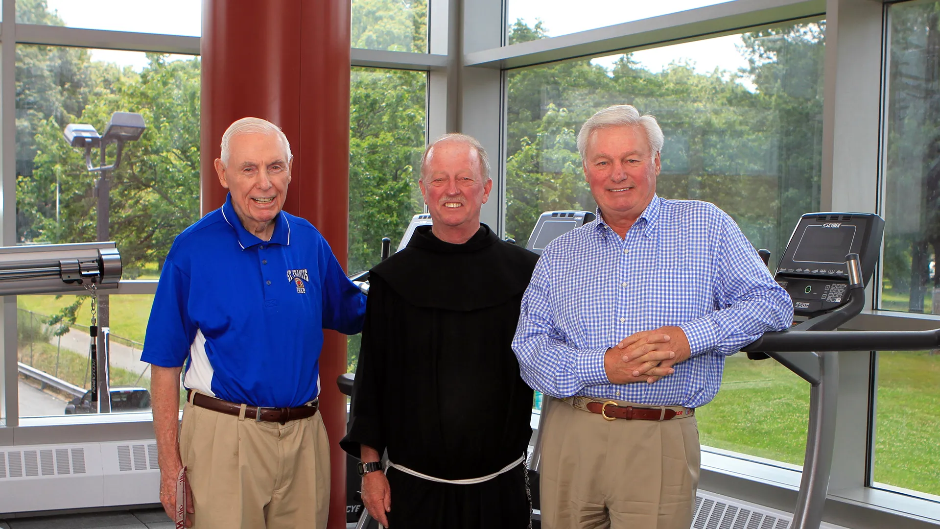 Color photograph of three older men standing together in a gym or fitness center with treadmills behind them, smiling at the camera, one wearing a religious habit and the others in casual shirts and slacks.