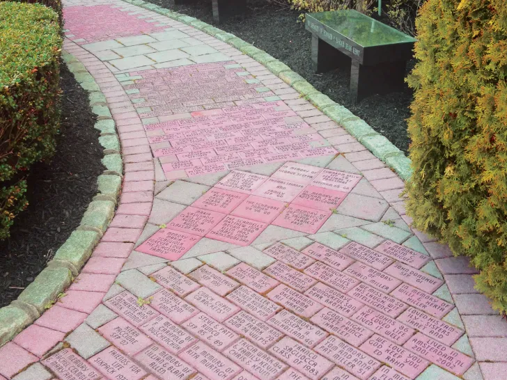 Curved brick memorial walkway with engraved dedication bricks, bordered by shrubs and featuring a stone bench in a landscaped garden.