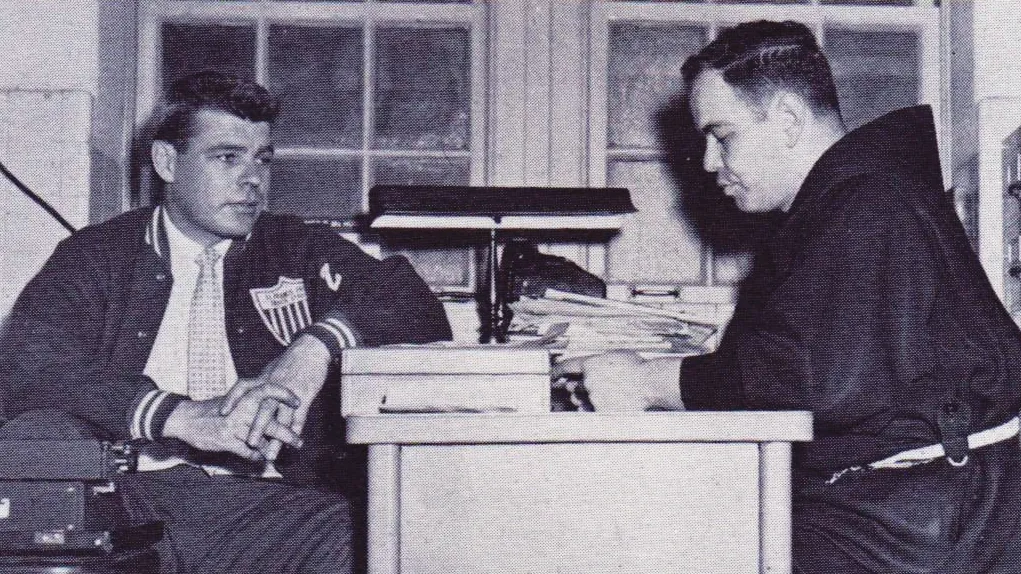 Black-and-white photograph of two men seated across from each other at a desk in an office or classroom, engaged in conversation, with books and papers stacked on the desk.
