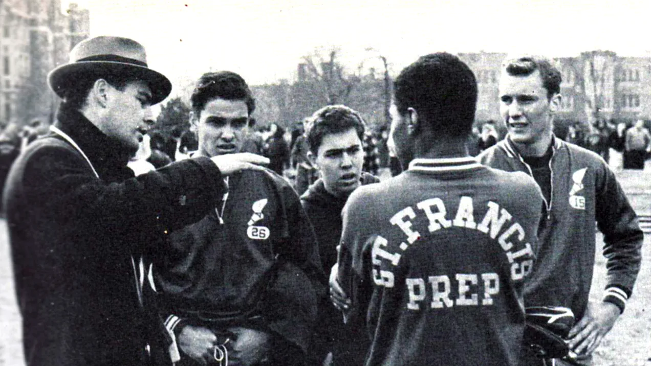 Black-and-white photograph of a priest or coach talking to a group of high school athletes wearing matching jackets on an outdoor field, with buildings visible in the background.