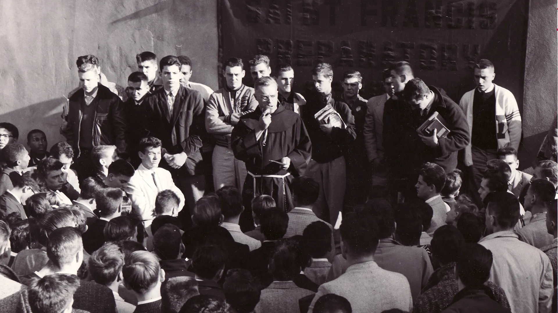 Black-and-white photograph of a Catholic friar speaking to a large crowd of young men gathered closely around him in an outdoor courtyard, many listening attentively.